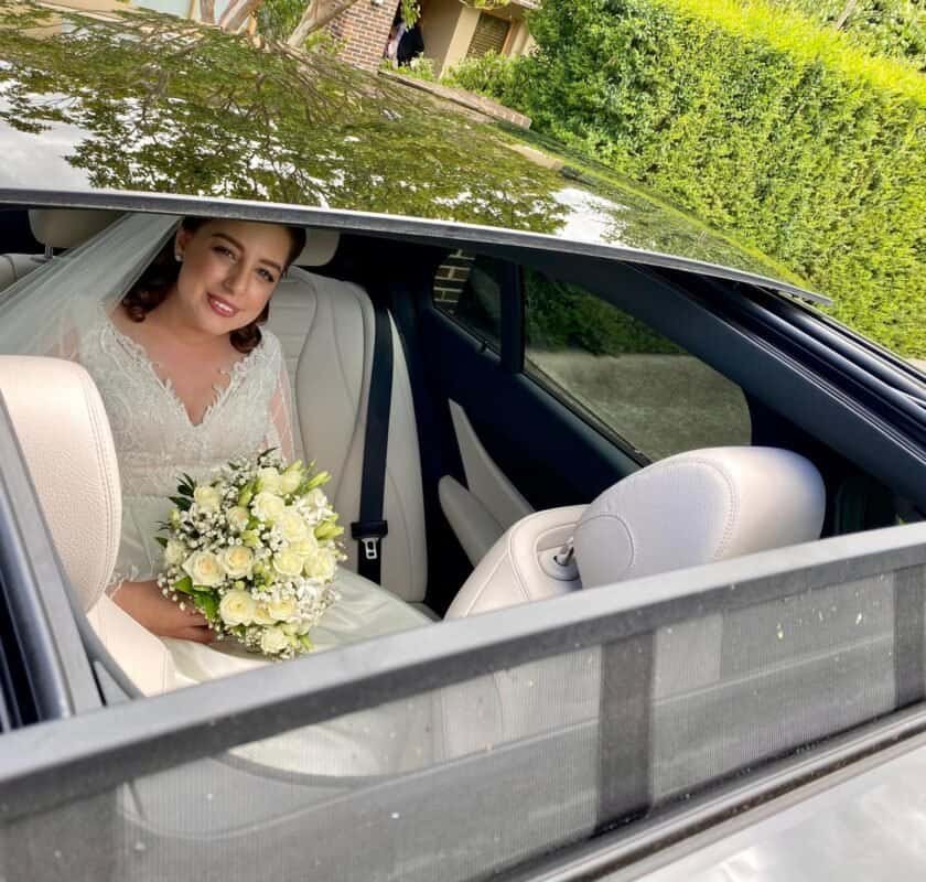 Bride in wedding dress holding a bouquet, sitting inside a sleek vehicle.
